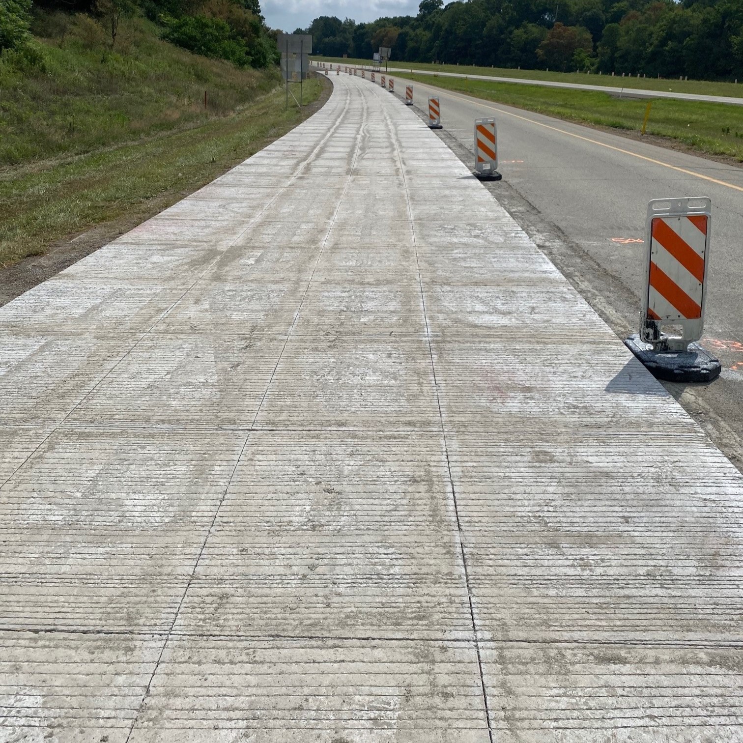 An image of a tree-lined, four-lane roadway with a grassy median between either side of the roadway with a line of orange and white work zone barricades between the lane where the thin-bonded concrete overlay was completed and the lane where it hasn't been completed yet  